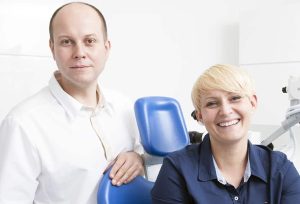 Dwie uśmiechnięte osoby w jasnym, białym gabinecie stomatologicznym w Krakowie. Jedna stoi i ma na sobie białą koszulę, druga siedzi w granatowej koszuli, a za nimi znajduje się sprzęt stomatologiczny - Profesjonalny Stomatolog Kraków.